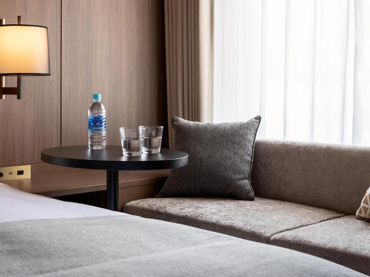 Mitsui Garden Hotel Roppongi Tokyo Premier Eksteriør billede The photo depicts a cozy hotel room setting. On the left, there is a bedside table with a lamp. Against the wall, there is a comfortable-looking couch with a decorative pillow. In front of the couch, there's a small round table that holds a bottle of