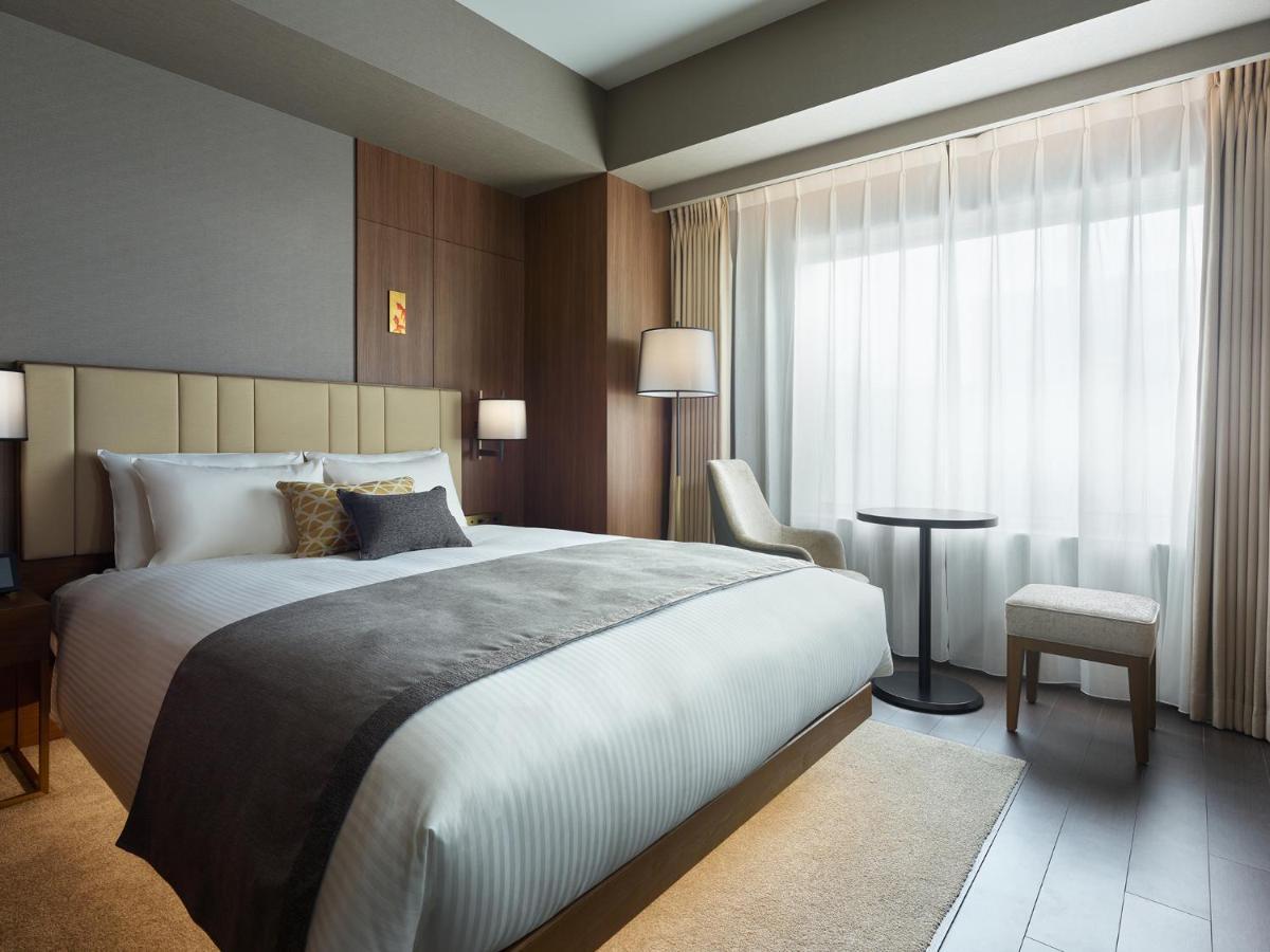 Mitsui Garden Hotel Roppongi Tokyo Premier Eksteriør billede The photo shows a modern hotel room featuring a comfortable bed with white linens and a decorative throw pillow. The room has a neutral color palette with wooden accents on the walls. There are two bedside lamps and a chair positioned next to a small