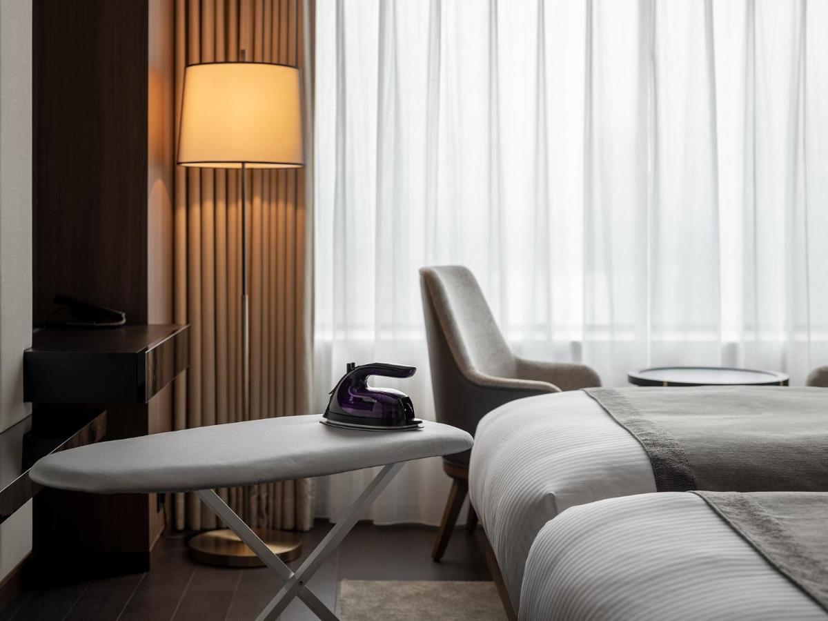 Mitsui Garden Hotel Roppongi Tokyo Premier Eksteriør billede The photo shows a modern hotel room featuring two beds with white bedding. There is a light-colored armchair next to a small side table. On the table, there is an iron, and beside it, a lamp with a warm glow is illuminating the space. The large windo