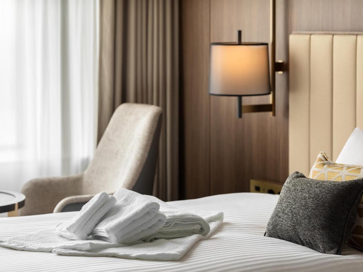 Mitsui Garden Hotel Roppongi Tokyo Premier Eksteriør billede The image shows a stylish hotel room. In the foreground, there is a neatly made bed with white linens, featuring a stack of white towels arranged in a decorative manner. To the side, there is a gray upholstered armchair and a small table. The backgro