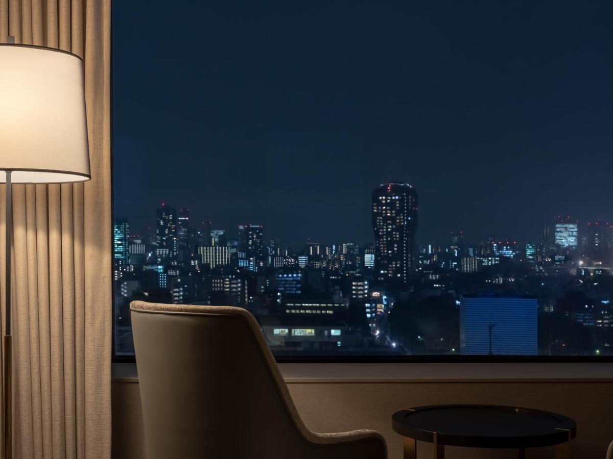 Mitsui Garden Hotel Roppongi Tokyo Premier Eksteriør billede The photo depicts a cozy hotel room setting at night. In the foreground, there's a stylish armchair next to a small round table. A standing lamp provides soft lighting, creating a warm ambiance. In the background, large windows reveal a city skyline 