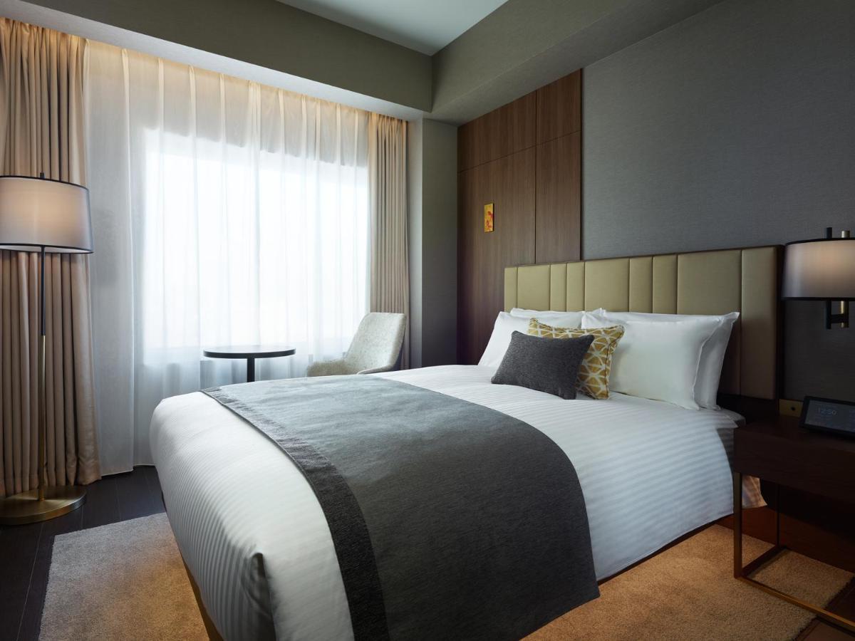 Mitsui Garden Hotel Roppongi Tokyo Premier Eksteriør billede The photo shows a modern hotel room featuring a neatly made bed with white linens and a decorative gray throw blanket. To the side, there is a small round table and a comfortable chair, creating a cozy seating area. The room is well-lit with natural 