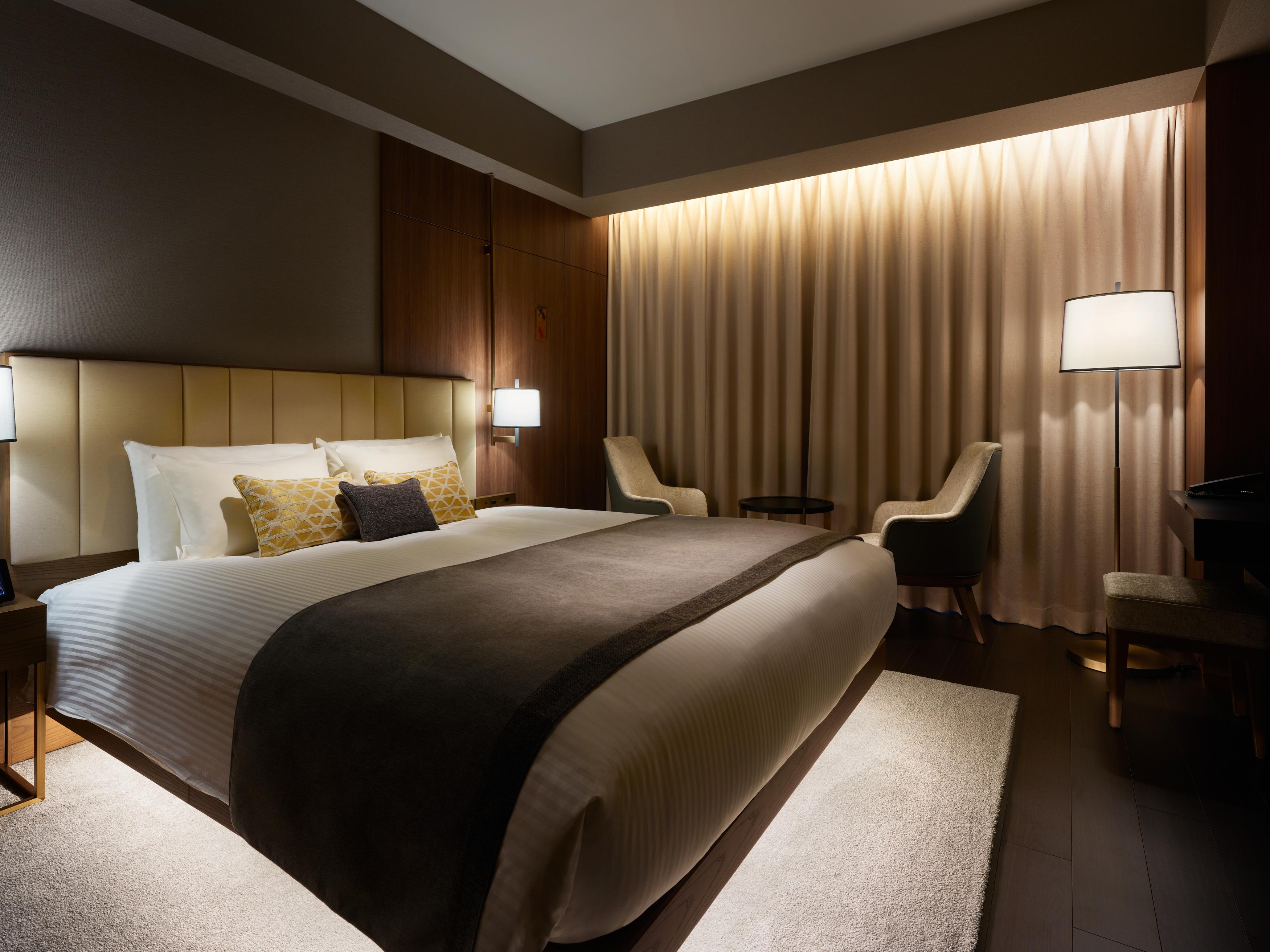 Mitsui Garden Hotel Roppongi Tokyo Premier Eksteriør billede The photo depicts a modern hotel bedroom. It features a large bed with white linens and a dark gray throw blanket, accented by decorative pillows. The room has a warm ambiance created by soft lighting, including two bedside lamps and a lighted area b