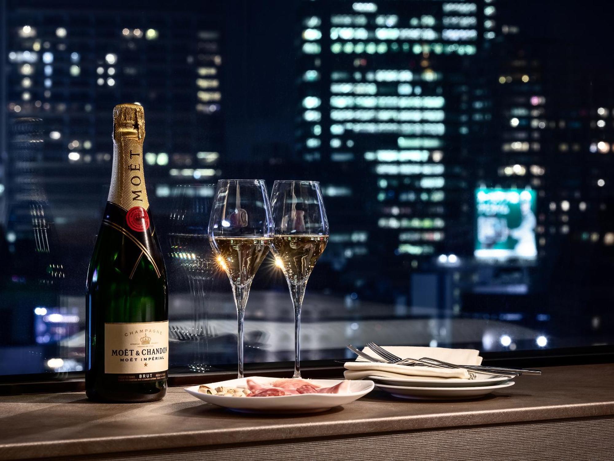 Mitsui Garden Hotel Roppongi Tokyo Premier Eksteriør billede The photo features a bottle of champagne, specifically Moët & Chandon, alongside two filled champagne glasses. The table setting includes a small plate with a selection of food and neatly arranged utensils. In the background, there is a view of a cit