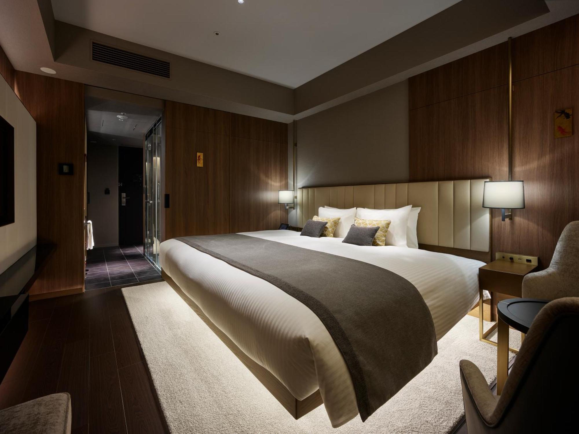 Mitsui Garden Hotel Roppongi Tokyo Premier Eksteriør billede The photo shows a modern hotel room featuring a large bed with a neutral-colored duvet and a dark throw blanket. The headboard is upholstered, and there are two bedside lamps on either side of the bed. The walls have a warm wood paneling, contributin