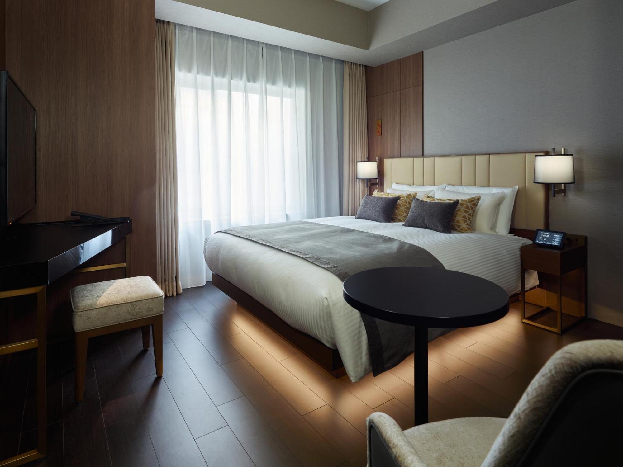 Mitsui Garden Hotel Roppongi Tokyo Premier Eksteriør billede The photo shows a modern hotel room featuring a large bed with decorative pillows. There are large windows with sheer curtains allowing natural light to filter in. To one side, there is a sleek desk with a chair and a small round table in the center 