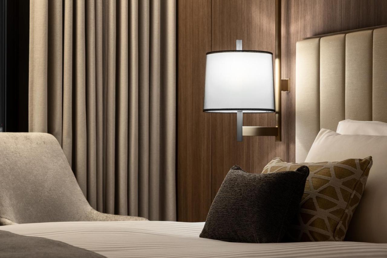 Mitsui Garden Hotel Roppongi Tokyo Premier Eksteriør billede The photo shows a cozy hotel bedroom setting. There is a neatly made bed with white linens and a few decorative pillows, including a dark one and another with a geometric pattern. To the side, there is a soft armchair and a modern wall lamp mounted a